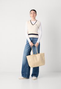 Beige Strickweste mit dunklen Streifen über einem weißen Langarmshirt, kombiniert mit weiten blauen Jeans und einer großen beigefarbenen Tragetasche.