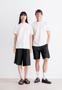 Young woman and man stand side by side wearing white t-shirts and black bottoms; woman in wide shorts with sandals, man in rolled shorts with loafers and socks.