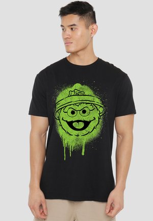 Black cotton t-shirt featuring a green graphic of a cartoon character’s face with splatter accents, emphasizing round eyes and a wide smile.