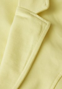 Lichtgele stof met een zachte textuur en subtiele ribbels. Voorzien van een opgevouwen kraag en nette stiksels langs de randen.