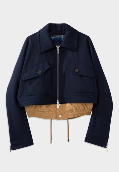 Chaqueta recortada de color navy con cuello puntiagudo, dos bolsillos en el pecho con cierres de botón y dobladillo y forro elásticos en color beige. Incluye cremalleras en los laterales.