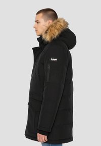 Sort dynejakke med hætte med pelskant, lynlåslomme på brystet og elastiske manchetter. Har et quiltet design og fremtrædende brandlogo.