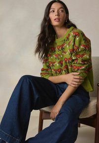 Green knitted sweater with a fox pattern, featuring short sleeves and ribbed neckline. Paired with wide-leg dark denim jeans.