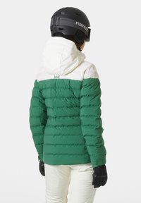 Groene pufjas met een witte bovensectie, voorzien van een capuchon, gestikt textuur en zwarte handschoenen. Draagt met witte skibroek.
