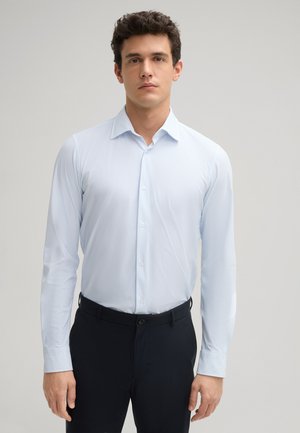 Helles blaues Langarmhemd mit feinem Muster, Knopfleiste und spitzem Kragen, kombiniert mit dunkler Hose.