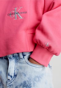 Felpa rosa con logo ricamato "Calvin Klein Jeans", polsini a costine, abbinata a pantaloni corti di jeans azzurri con effetto usurato. Tessuto liscio.
