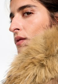 Cuello de piel sintética, color marrón claro con acentos en marrón claro, rodeando un rostro. Piel suave, rasgos definidos y maquillaje natural. Fondo brillante.