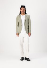 Blazer verde deschis cu două nasturi, tricou alb și pantaloni bej. Pantofii negri completează ținuta. Material neted, croi ajustat.