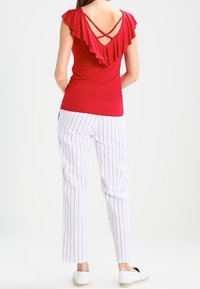 Rotes ärmelloses Top mit rüschigem Rücken und überkreuzten Trägern, kombiniert mit weißen Hosen mit blauen vertikalen Streifen und flachen Schuhen.