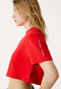 T-shirt cropped rossa realizzata in tessuto morbido, con collo rotondo e logo stampato "RED BULL RACING" sulla manica sinistra in bianco.