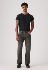 Must värvikas lühikeste varrukatega T-särk, millel on tasku, koos tumedate hallide sirgete pükstega ja mustade kingadega. Modell seisab käed taskutes.