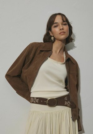 Braune Wildlederjacke mit Reißverschluss, cremefarbenes Tanktop, beige plissierter Rock und brauner Gürtel mit rundem Metallverschluss und geprägtem Muster.