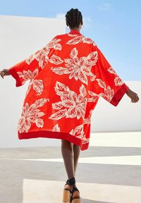 Kimono rouge avec de grandes fleurs blanches, manches larges et bordure contrastée à motifs à l'ourlet. Tissu léger avec une coupe ample.