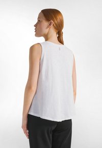 Top blanco sin mangas con un corte relajado y dobladillo redondeado, con una textura sutil y una pequeña etiqueta en la parte posterior del cuello. Combinado con pantalones negros.