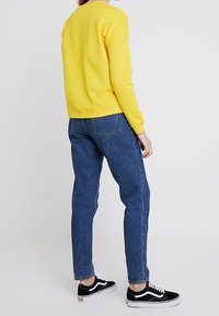 Felpa gialla con scollo rotondo, jeans blu a gamba stretta e sneaker nere con suole e dettagli bianchi.