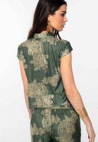 Camisa de manga corta con patrón verde y dorado, de tela texturizada y con cuello; presenta una silueta recortada y una abertura en la parte trasera.