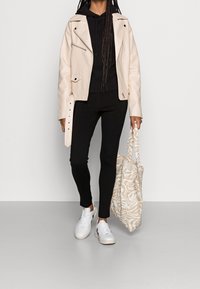 Jasnobeżowa kurtka motocyklowa z eko skóry ze srebrnym suwakiem, czarną bluzą z kapturem, czarnymi legginsami, białymi trampkami i beżową torbą na ramię w wzory.