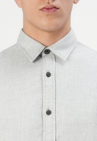 Camisa de botón gris claro con una tela texturizada, que presenta un collar puntiagudo y botones oscuros a lo largo del placket frontal.