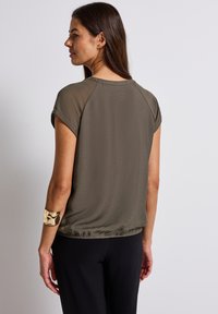 Top verde a maniche corte caratterizzato da una texture liscia, vestibilità rilassata e orlo arricciato. Accentuato da un elegante bracciale dorato al braccio.