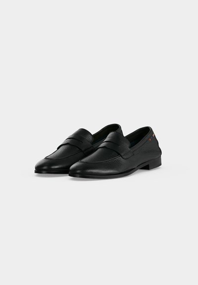 Paire de mocassins en cuir noir avec un petit talon et une bride sur le dessus, présentée sur un fond clair uni.