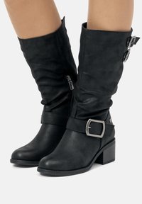 Botas negras hasta la mitad de la pantorrilla con tacones cuadrados, cremallera lateral y hebillas de metal decorativas en las correas alrededor de los tobillos y la parte superior de los ejes.