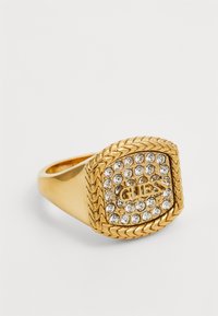 Goldring mit quadratischem Kopf, versehen mit einem strukturierten geflochtenen Rand und besetzt mit klaren Kristallen. In der Mitte steht das geprägt Wort "GUESS."