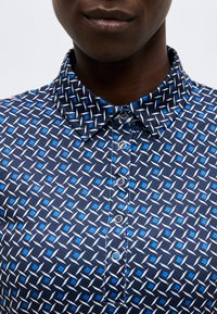 Κοντινό πλάνο ενός ατόμου που φοράει μπλε πουκάμισο με άσπρα και σκουρομπλε γεωμετρικά σχέδια και ασημένια κουμπιά με πατινέτα.