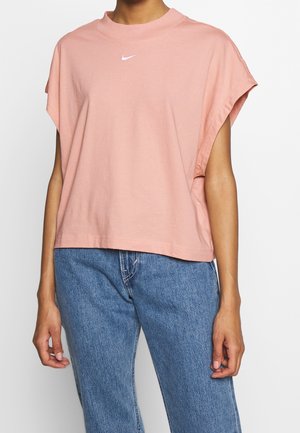 Femme portant un débardeur rose ample Nike et un jean bleu, visible du cou aux hanches sur un fond blanc.