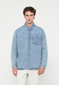 Camisa de denim azul claro com fecho de botões, gola clássica e bolso na parte esquerda do peito com um padrão subtil. Mangas longas com punhos.