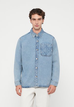 Lichtblauw denim overhemd met knoopsluiting, klassieke kraag en een linkerborstzak met een subtiel patroon. Lange mouwen met manchetten.
