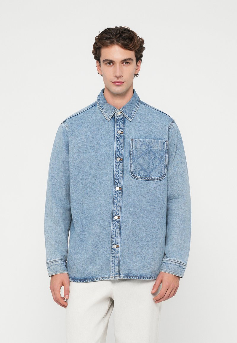 Camisa de denim azul claro com fecho de botões, gola clássica e bolso na parte esquerda do peito com um padrão subtil. Mangas longas com punhos.