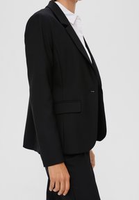 Czarna dopasowana marynarka z klapami, z jednorzędowym zapięciem na guzik i dwoma bocznymi kieszeniami. Wykonana z gładkiego materiału o usztywnionym kroju.