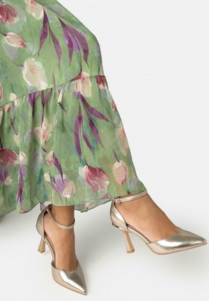 Piernas con tacones de tiras en el tobillo de color dorado metálico combinados con una falda verde fluida con patrones florales en púrpura y rosa.