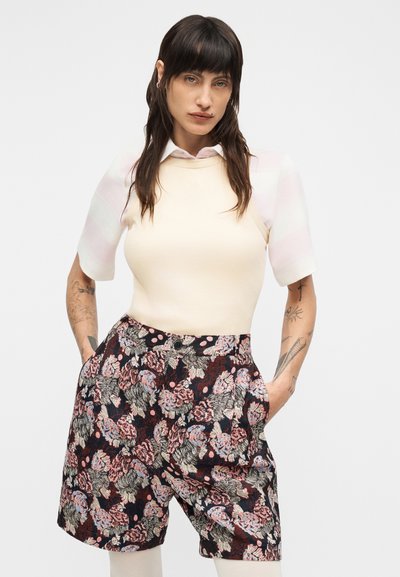 Ελαφρύ κρεμ εφαρμοστό τοπ με κοντά ριγωτά μανίκια, σε συνδυασμό με μαύρο σορτς με λουλουδάτο μοτίβο, τσέπες και εμφανείς λεπτομέρειες κουμπιών.