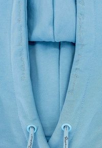Felpa azzurra chiara con una texture liscia, dotata di cordino. I dettagli ricamati lungo i bordi aggiungono un sottile interesse di design.