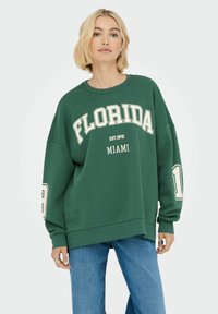 Felpa verde con la scritta "FLORIDA" e "MIAMI" stampata in bianco, con vestibilità over-size, maniche lunghe e polsini a costine. Indossata con jeans blu.