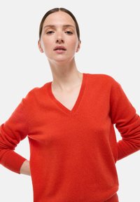Femme à la peau claire et aux cheveux châtain clair tirés en arrière, portant un pull V orange vif, se tenant sur un fond clair uni.