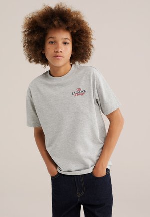 Jeune personne aux cheveux bouclés portant un t-shirt gris avec un texte rouge et noir et un jean foncé, mains dans les poches, debout devant un fond uni.