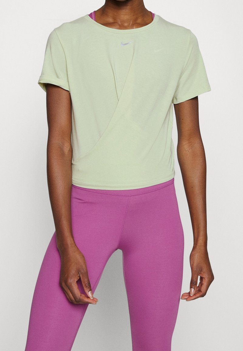 Hellgrünes Nike T-Shirt mit kurzen Ärmeln und einem Kreuzdesign auf der Vorderseite, kombiniert mit pinken Leggings. Weicher Stoff, lockere Passform.