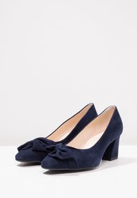 Scarpe con tacco alto in suede blu navy con punta rotonda e un dettaglio di fiocco decorativo nella parte anteriore. Presentano un tacco largo per una maggiore stabilità.