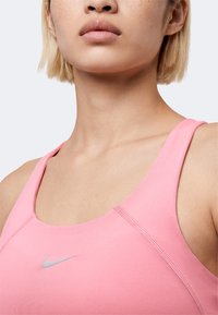 Κοντινό πλάνο ατόμου που φοράει ροζ αθλητικό σουτιέν Nike, δείχνοντας τον λαιμό, τους ώμους και το πάνω μέρος του στήθους με ορατό λογότυπο.