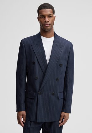 Mann trägt dunkelblauen Nadelstreifen-Doppelreiher-Blazer über weißem Hemd und steht vor einem schlichten hellgrauen Hintergrund.
