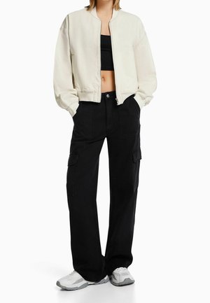 Krémová bomber bunda, černý croptop, černé cargo kalhoty a bílé tenisky. Bunda má zip a žebrované manžety; kalhoty mají velké kapsy.