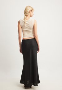 Ženska s blond kitkami nosi bezrokavni bež top, dolga črna krila in črne petke, stoji in se obrača stran na enotni ozadju.