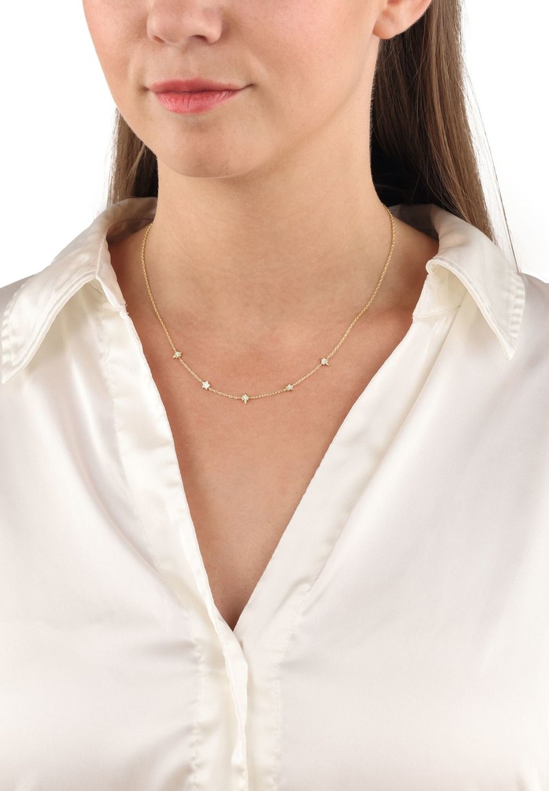 Guldtonad halsband med en delikat kedja som har fem stjärnformade detaljer, buren över en vit satinskjorta.