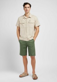 Beige kortärmad skjorta med två bröstfickor, gröna shorts och bruna sandaler. Avslappnad passform, lätt tyg, platt krage och ren design.