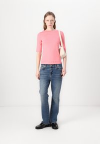 Roze getailleerde top met driekwart mouwen; blauwe ontspannen jeans; zwarte schoenen; model houdt een kleine lichtgekleurde handtas vast.
