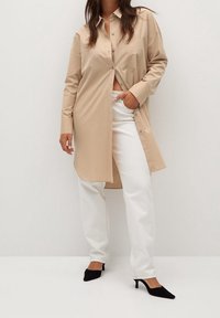 Chemise beige à boutons, manches longues, poche avant, associée à un pantalon blanc. Talons noirs à bout pointu. Textures de tissu simples et lisses.