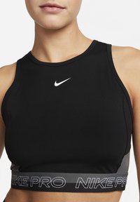 Sort atletisk crop top med en tætsiddende design, rund halsudskæring og et hvidt Nike-logo på brystet. Den har et gråt bånd med teksten "NIKE PRO".