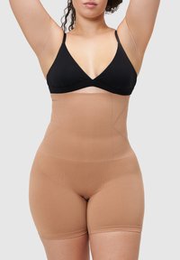 Beżowy, wysoki pas gorsetu modelującego wykonany z gładkiej, elastycznej tkaniny. Posiada bezszwową konstrukcję z cięciem na wysokości środkowej części uda i bez widocznych zapięć.
